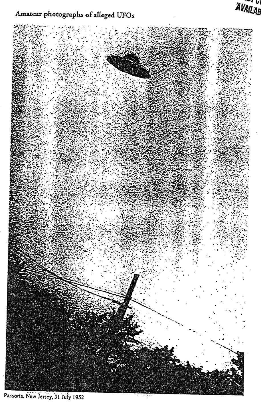 Fotografia granulada em preto e branco de um objeto escuro em forma de disco no céu, acima de fios elétricos, de um poste inclinado e de arbustos.