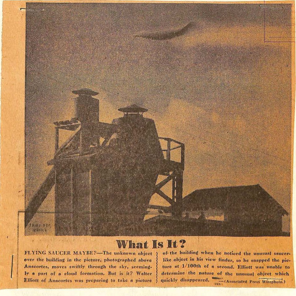 Fotografia de jornal em tons de sépia de uma torre industrial e um galpão; no alto, à direita do centro, uma mancha escura alongada no céu.