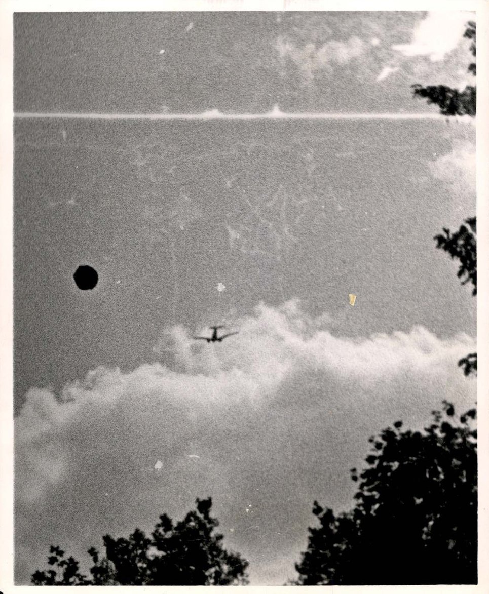 Fotografia em preto e branco do céu com um avião bimotor ao centro e um ponto escuro redondo à sua esquerda, com copas de árvores nas bordas.