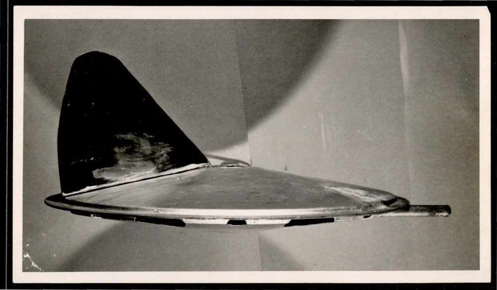 Fotografia em preto e branco de um disco metálico achatado, visto de lado, com uma aleta triangular escura.