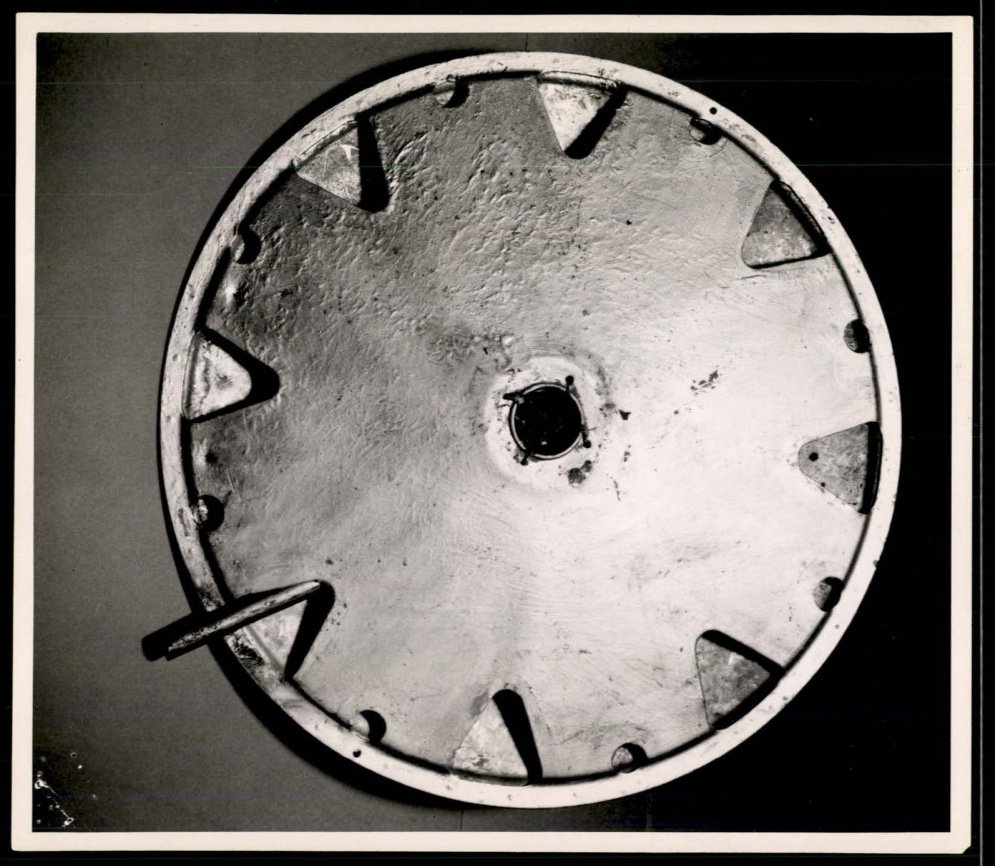 Fotografia em preto e branco da face inferior de um disco metálico com furo no centro e recortes triangulares na borda.