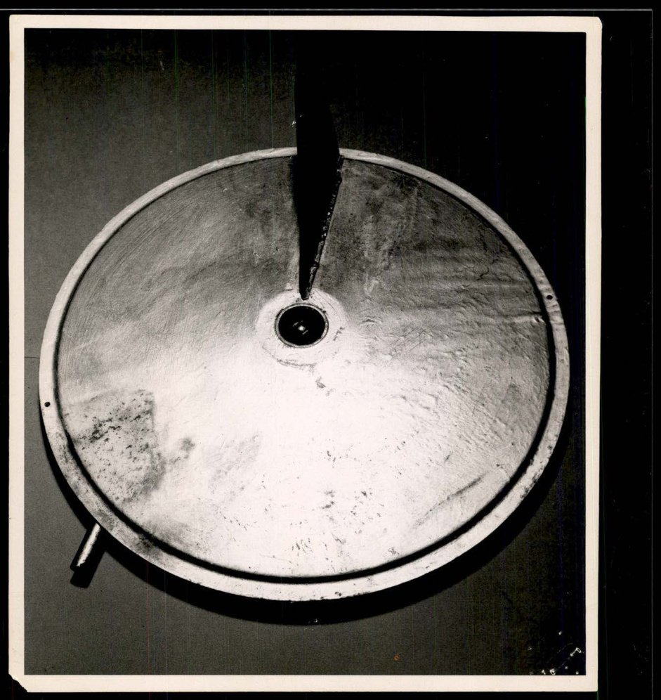 Fotografia em preto e branco de um disco metálico visto de cima, com uma aleta escura e um furo no centro.