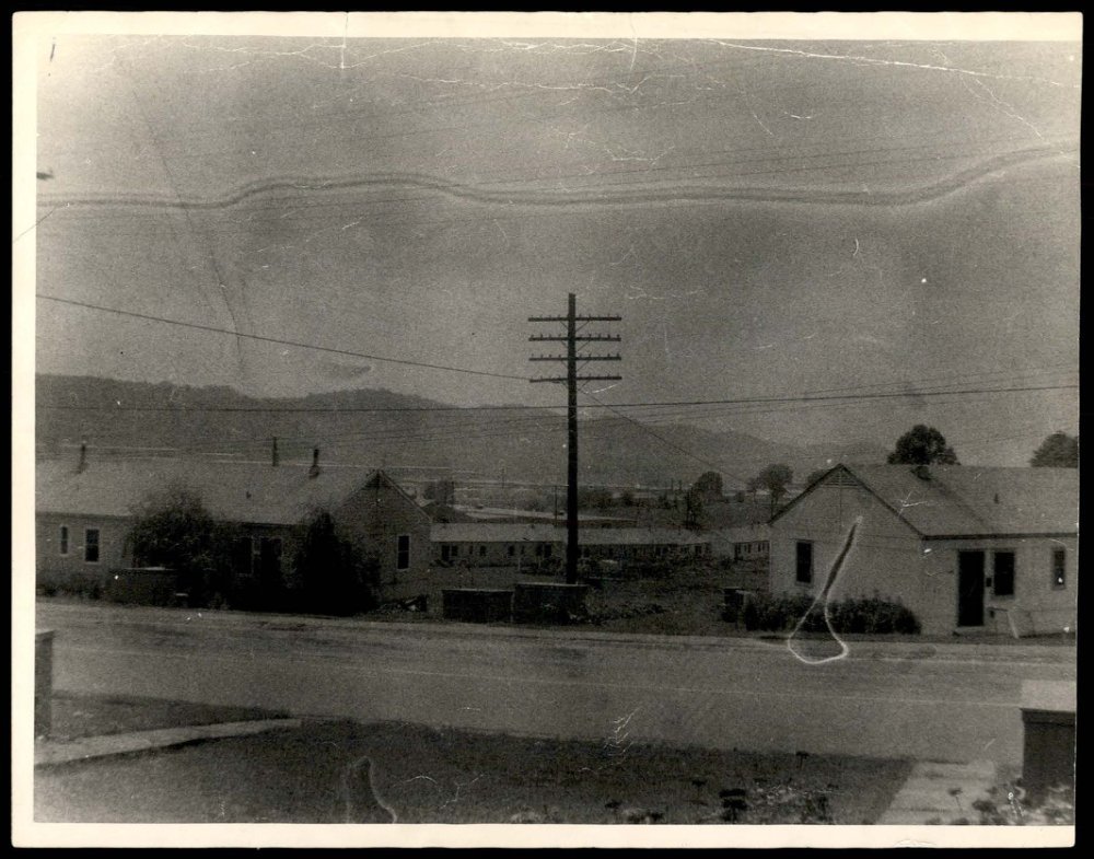Foto em preto e branco de uma rua com casas térreas, um poste de madeira e montanhas ao fundo; uma linha clara ondulada cruza o céu.