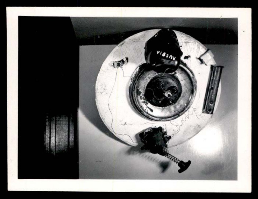 Fotografia em preto e branco de um disco artesanal com fios e peças mecânicas e uma caixa com a palavra RUSSIA.
