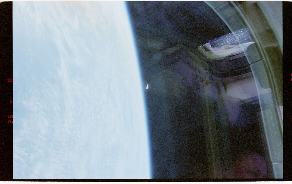 Borda da Terra à esquerda e espaço escuro à direita; pequeno objeto branco triangular perto do centro; reflexos da cabine no vidro.